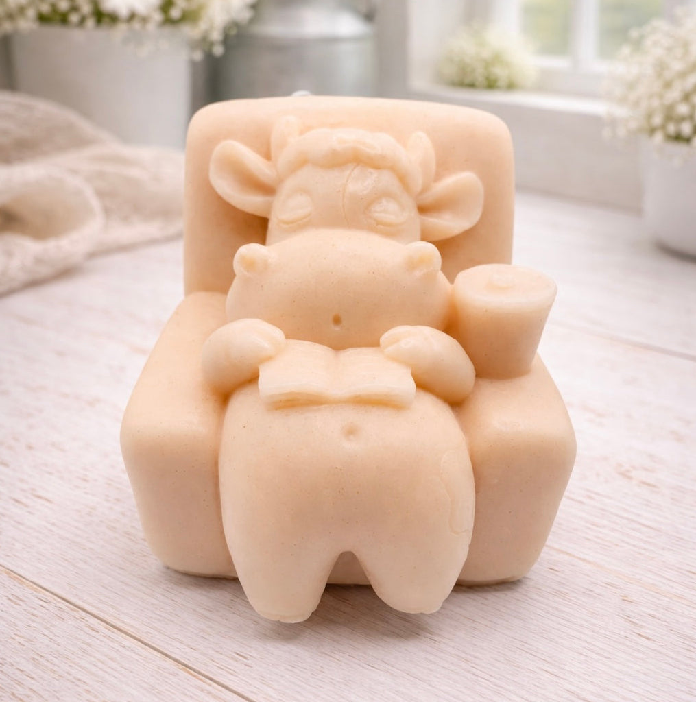 A handmade vegan soap sculpted to show a whimsical "lazy cow" character relaxing in an armchair, reading a book and holding a drink, displayed on a light wooden surface.