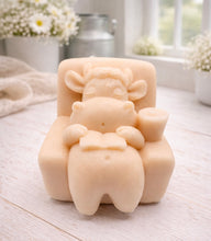 Load image into Gallery viewer, A handmade vegan soap sculpted to show a whimsical "lazy cow" character relaxing in an armchair, reading a book and holding a drink, displayed on a light wooden surface.

