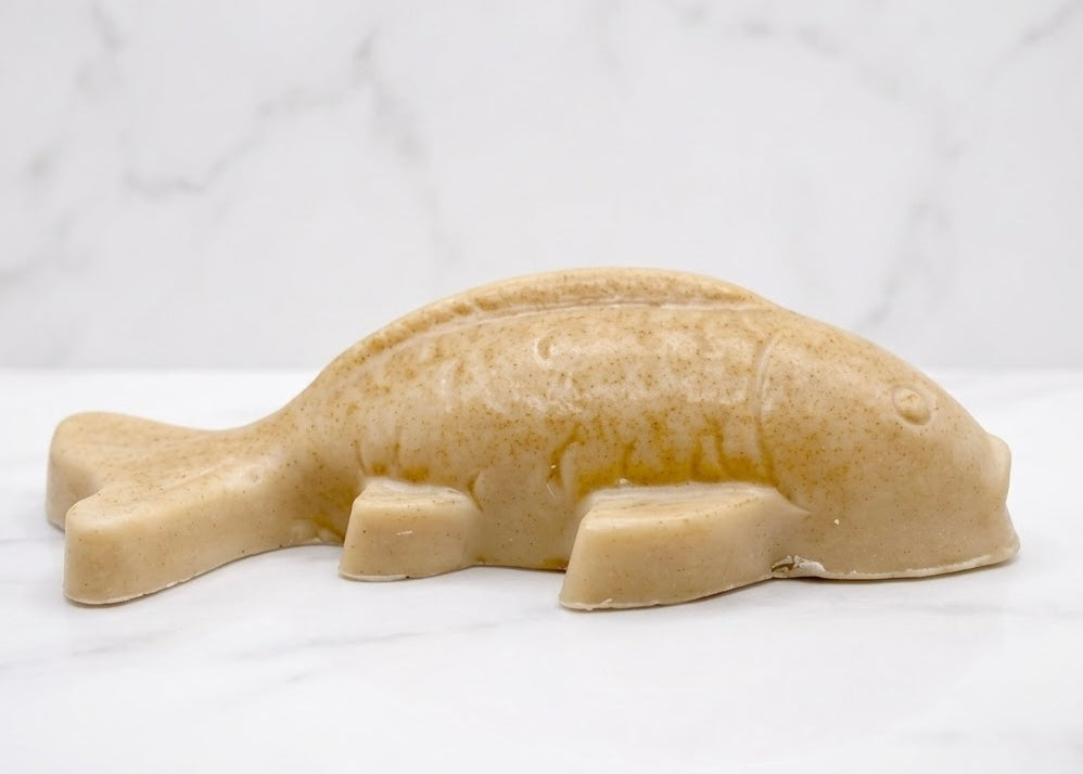 Handmade yellow vegan soap sculpted in the shape of a koi carp fish with detailed scales and fins, resting on a light timber surface with a soft floral background.