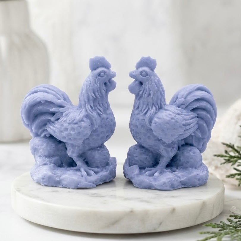 Two intricate, pastel blue cockerel soaps stand on a round white marble trivet, set in a neutral bathroom. Each soap is moulded with fine wattle and tail feather details, creating a charming, miniature bird sculpture in a calming lavender hue.
