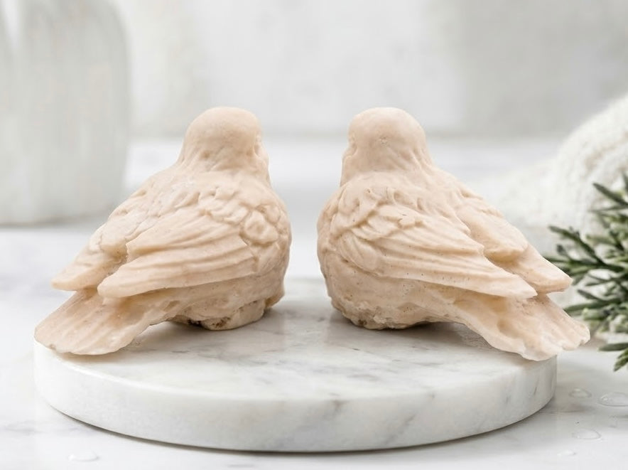 A rear view pair of two bird soaps in a light yellow tone, displayed side by side on a round white marble board against a softly blurred light background.
