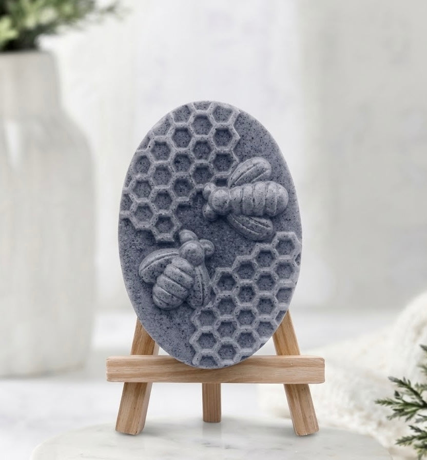 A close-up photograph of an oval, dark grey charcoal soap bar with raised carvings of two bees and honeycomb details. It is presented on a mini wooden display easel resting on a marble base.