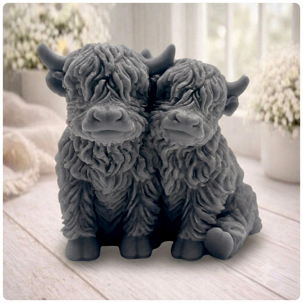 Handmade soap featuring two Highland cows with textured, shaggy fur sitting side by side on a light wooden surface with a softly indoor background.