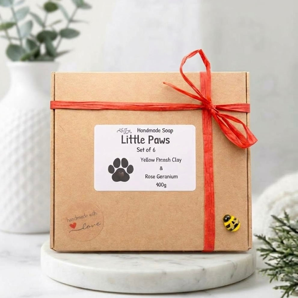 A brown cardboard box gift set containing six 'Little Paws' handmade soaps. The box is tied with red ribbon, has a decorative paw print label, and sits on a marble stand with greenery.