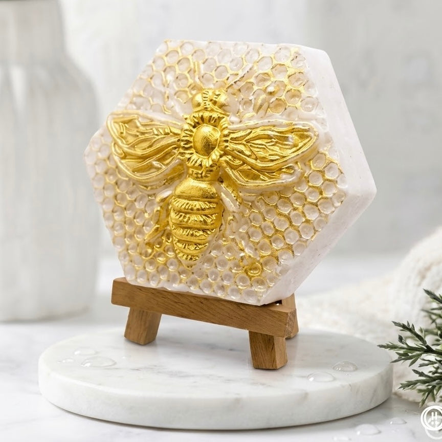 A hexagonal honeycomb-textured soap, now seen from a three-quarter angle from the left, highlighting the raised gold bee relief and the detailed hexagonal pattern. The soap is still displayed on its wooden easel