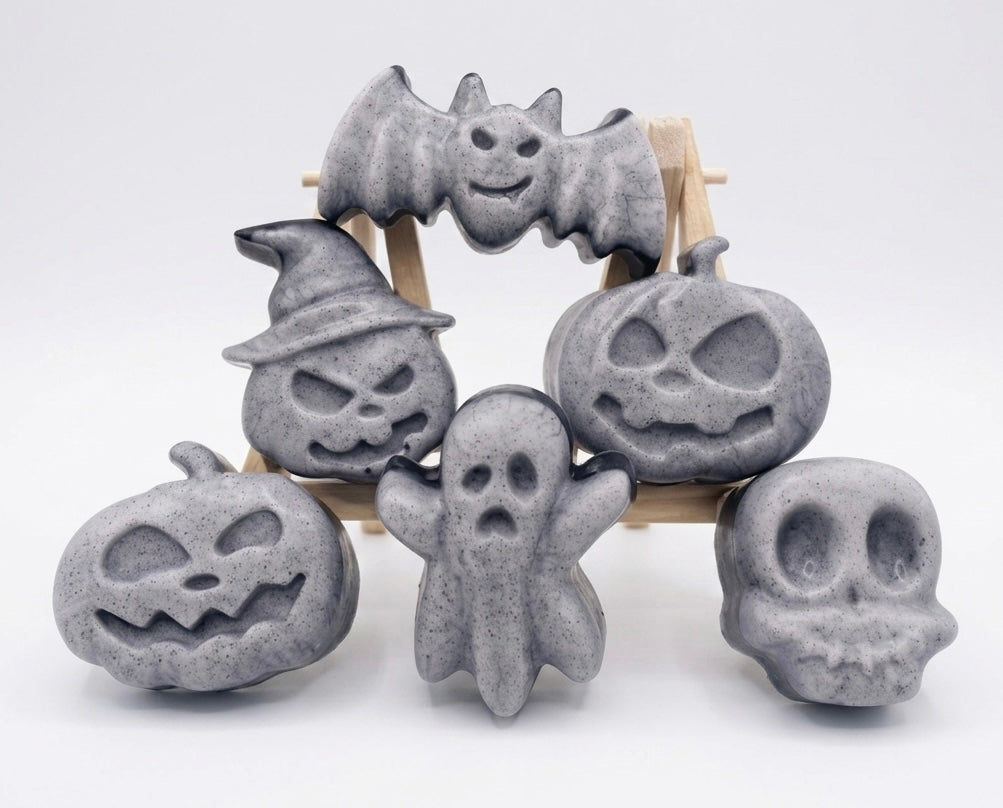 A collection of six handmade vegan Halloween soaps in a mottled grey and black charcoal finish, featuring a smiling bat, three different carved pumpkins (one wearing a witch's hat), a ghost, and a skull, arranged on and around a small wooden easel.