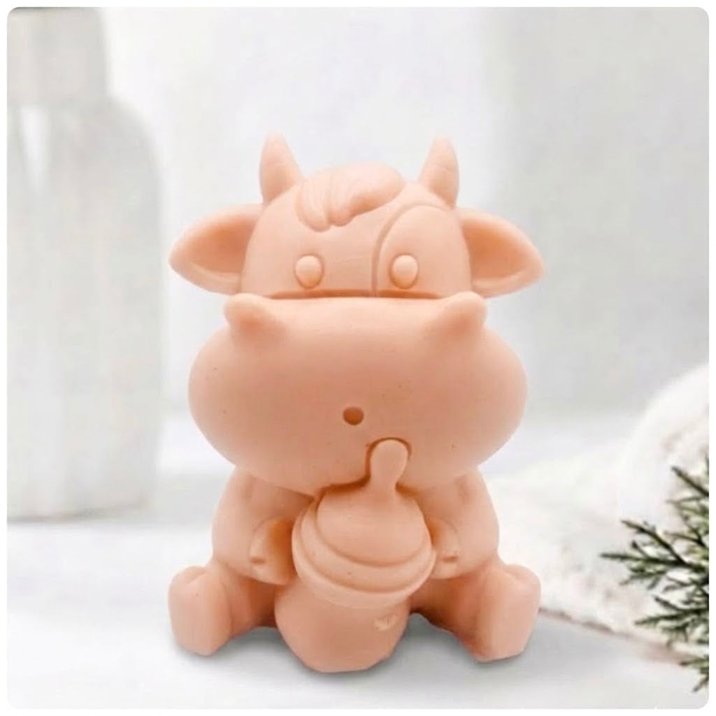 A baby cow-shaped soap holding a small milk bottle, photographed on a soft white background with a blurred bottle and greenery in the surroundings.
