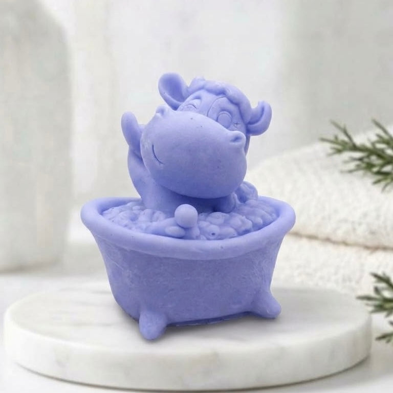 Novelty vegan soap gift sculpted into a cute lavender blue cartoon cow happily waving while sitting inside a miniature footed bathtub filled with soap bubbles.