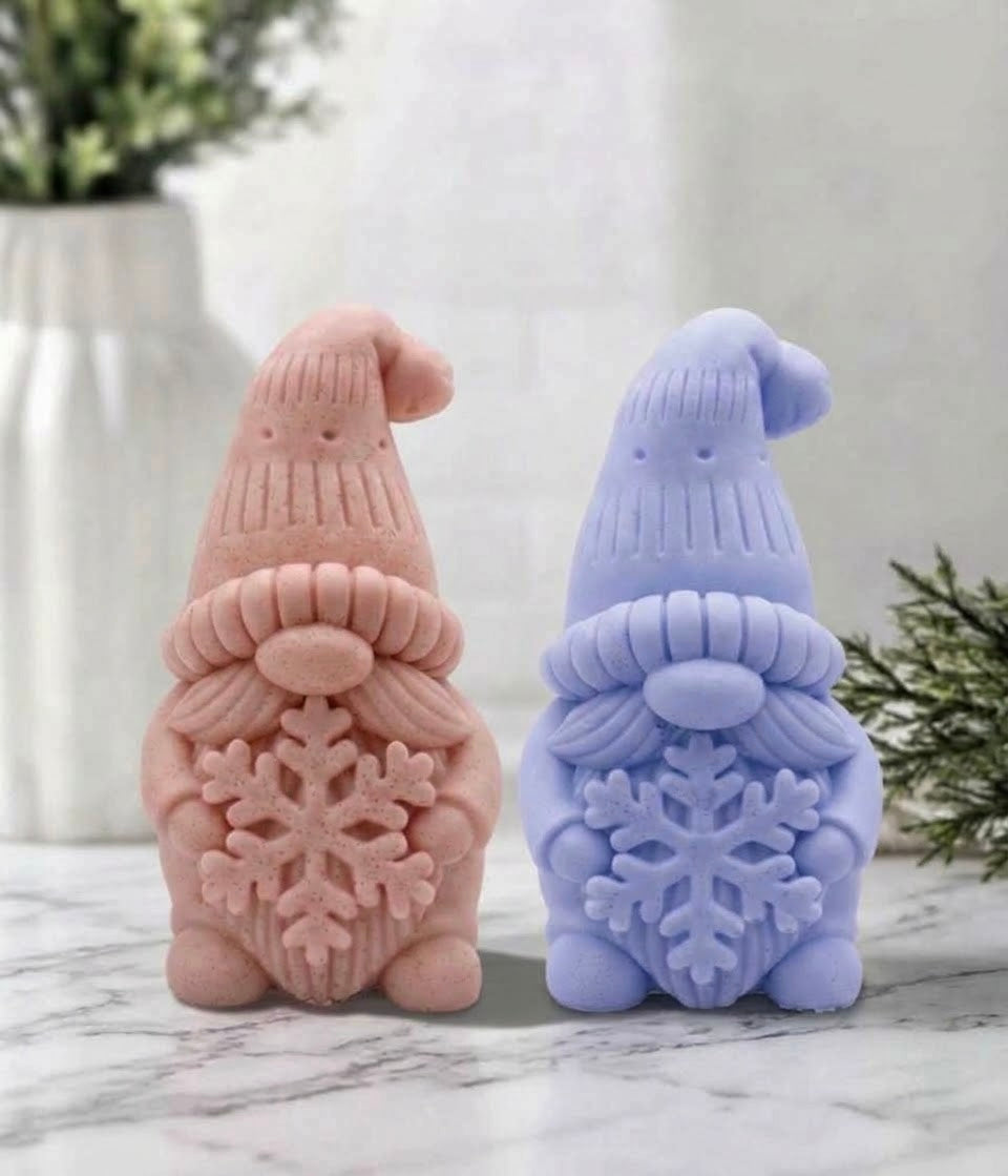 Two gnome-shaped soap bars standing side by side on a marble surface. The left one is a dusty pink, and the right one is a periwinkle blue, both featuring textured knit hats, beards with snowflake details, and small hats on top.
