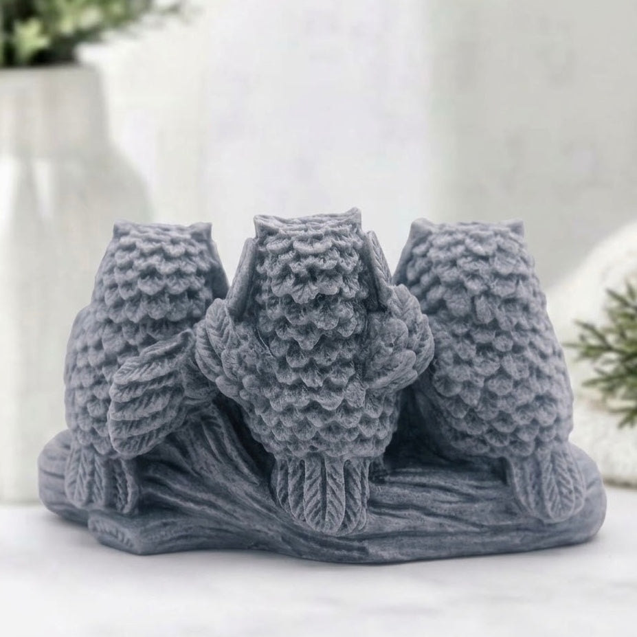 A grey decorative soap of three owls perched on a branch, photographed on a light surface with a softly blurred indoor background.