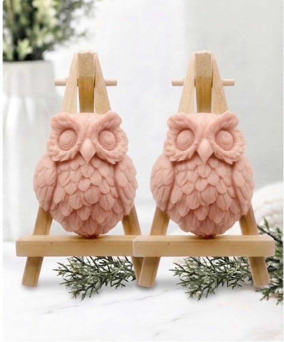 Two owl-shaped handmade soaps in soft, dusty pink displayed side by side on small wooden easel stands on a white marble surface.
