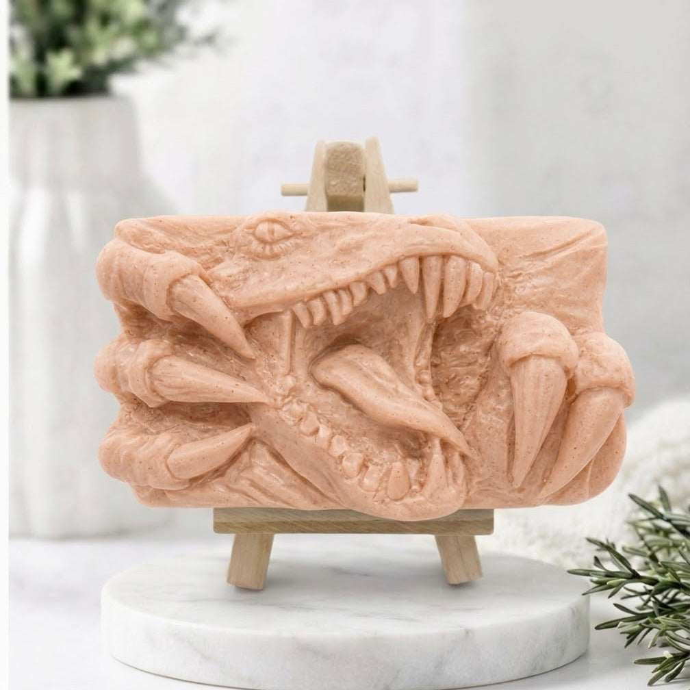 A rectangular soft pink handmade vegan soap  featuring a high-relief carving of a Tyrannosaurus Rex with its jaws wide open in a roar and sharp claws framing the face, displayed on a miniature wooden easel.