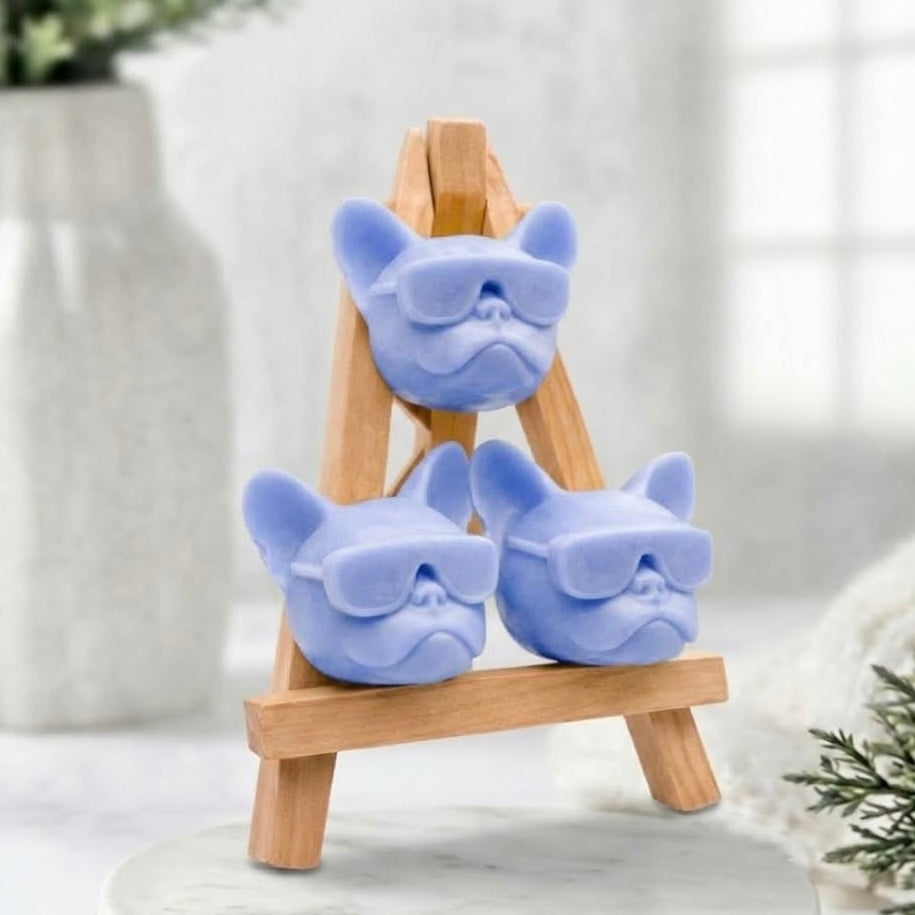 Handmade French Bulldog novelty soap gift set. Three artisan, soft blue Frenchie-head soaps wearing matching sunglasses are stacked and displayed on a natural wooden artist's easel.