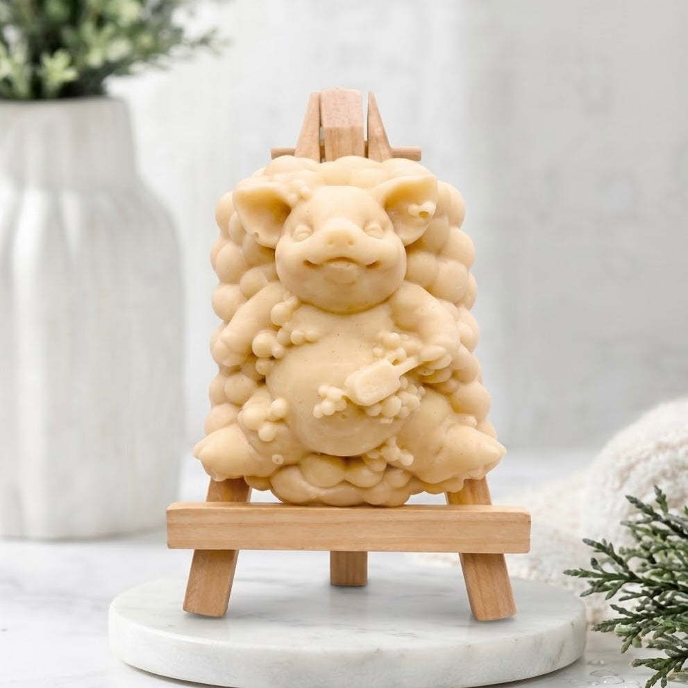 A product shot of a pig-shaped soap figurine with a bubble-cluster base, made in a pale yellow colour, positioned full-frontal on a miniature wooden easel in a simple setting.