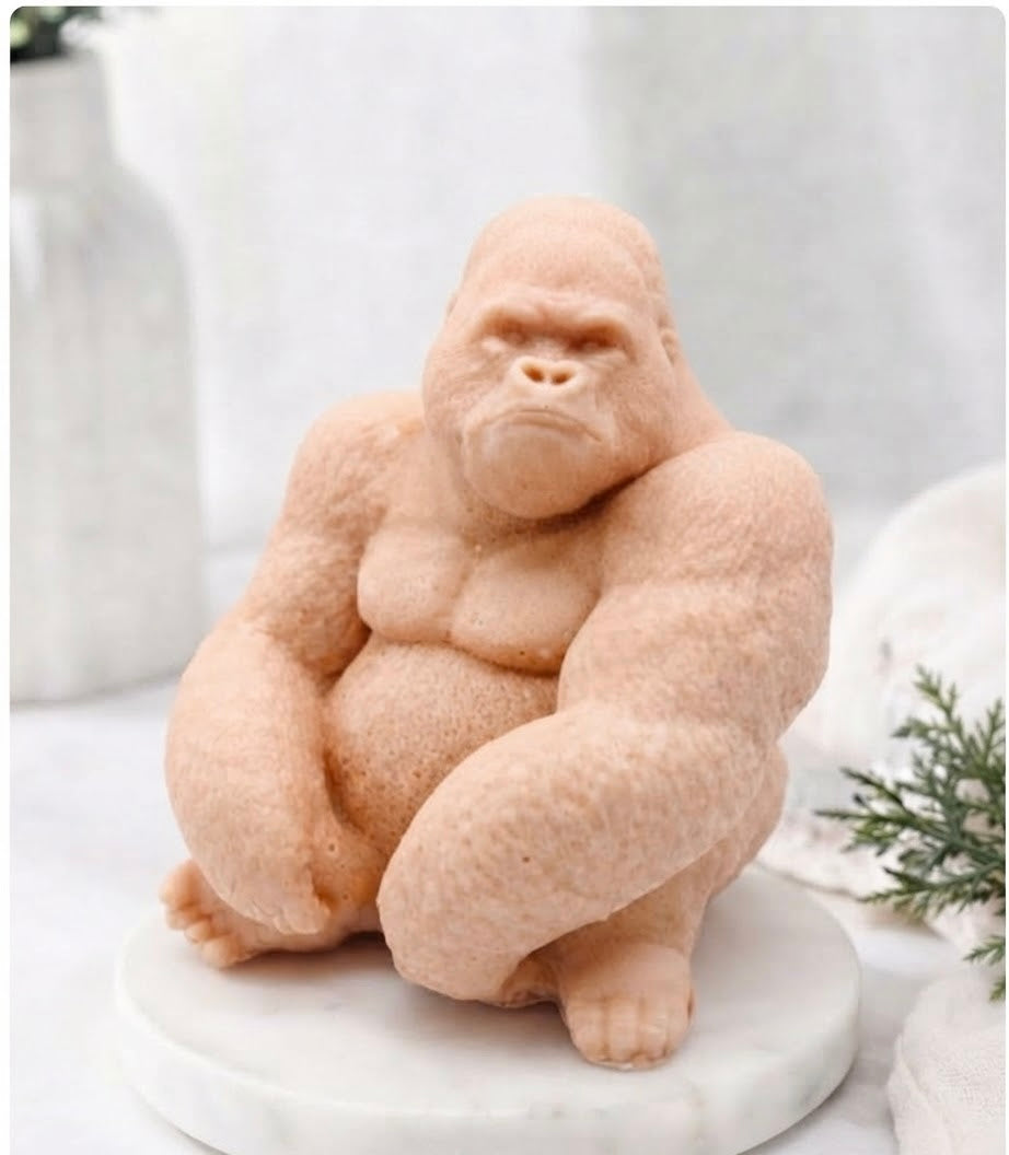 A sculpted gorilla soft pink coloured vegan soap in a seated pose on a round white marble-like base, photographed in a softly styled indoor setting with blurred decor in the background.