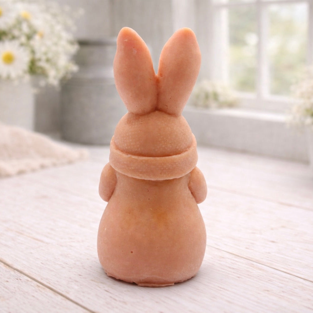 A detailed handmade vegan soap in a natural colour, sculpted as a whimsical Easter gnome (gonk) with long rabbit ears and a bow on its hat, holding a patterned Easter egg, displayed on a light wooden surface. Easter gift 