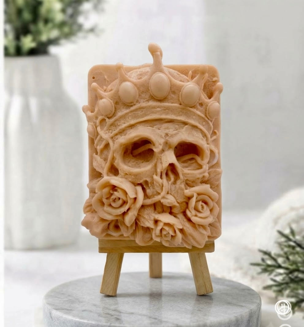 A detailed photograph of an artisanal red French clay vegan soap in the shape of a crowned human skull with detailed rose flowers, displayed on a wooden easel against a soft, bright marble and home decor background.