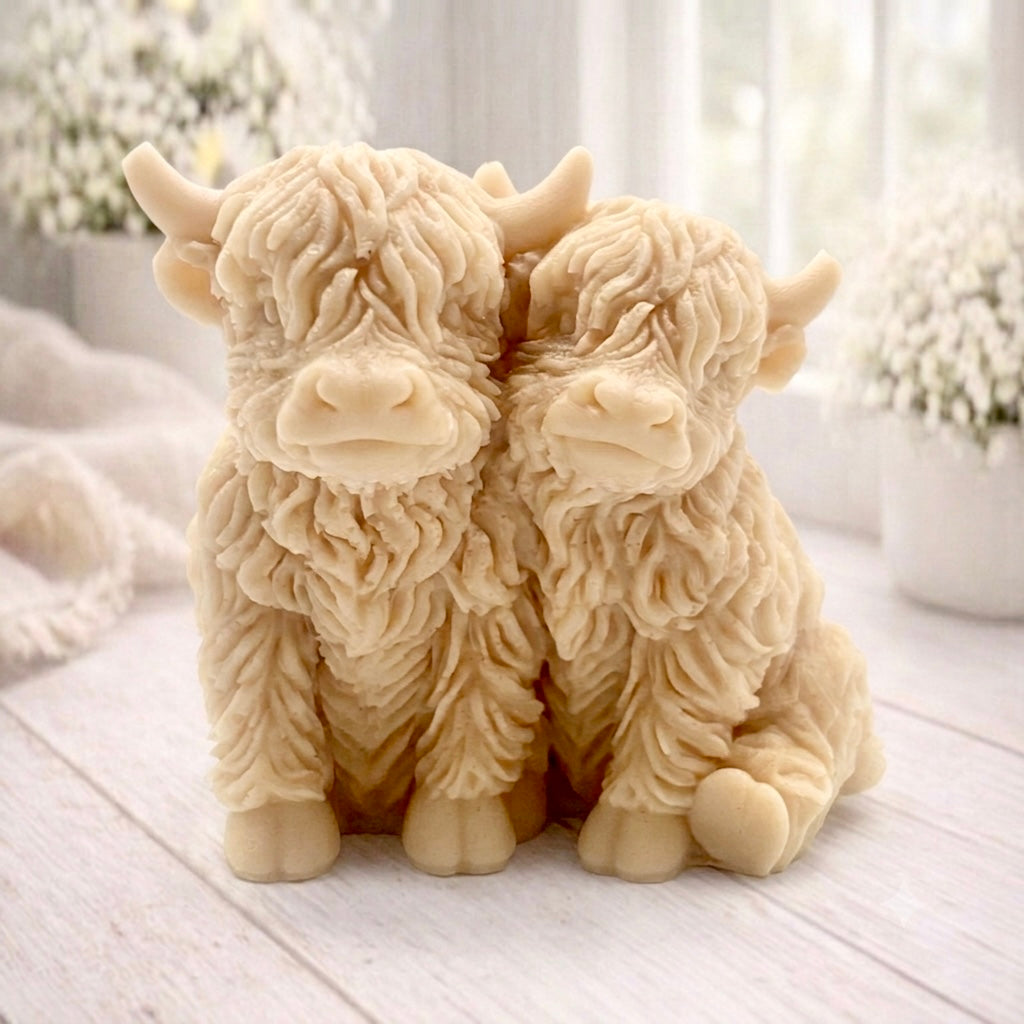 Handmade soap featuring two Highland cows with textured, shaggy fur sitting side by side on a light wooden surface with a softly indoor background.