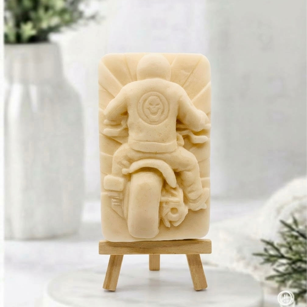 A Yellow-coloured natural handmade motorbike soap bar with carved rider and skull patch design, displayed on a wooden easel stand