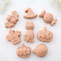 A soft pink collection of eight handmade vegan Halloween soaps featuring a bat, witch's hat, star-patterned sweet, haunted house, black cat, jack-o'-lantern, and a ghost, arranged on a light wooden surface with white floral accents.