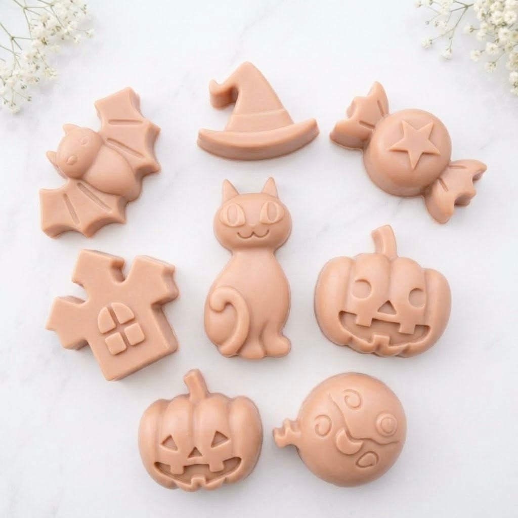 A soft pink collection of eight handmade vegan Halloween soaps featuring a bat, witch's hat, star-patterned sweet, haunted house, black cat, jack-o'-lantern, and a ghost, arranged on a light wooden surface with white floral accents.