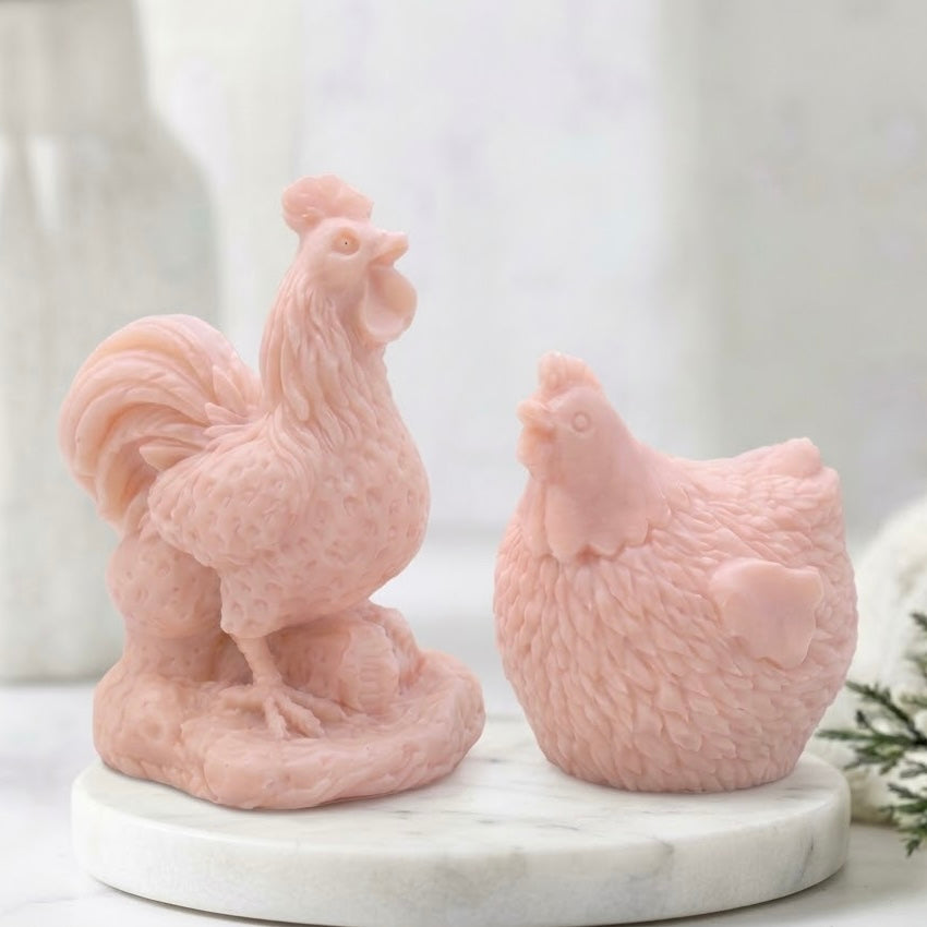 Side profile of two intricately detailed, pastel pink, vegan soap bars - one cockerel and one hen - sitting on a white marble coaster with a bokeh background of a window and blurry greenery.