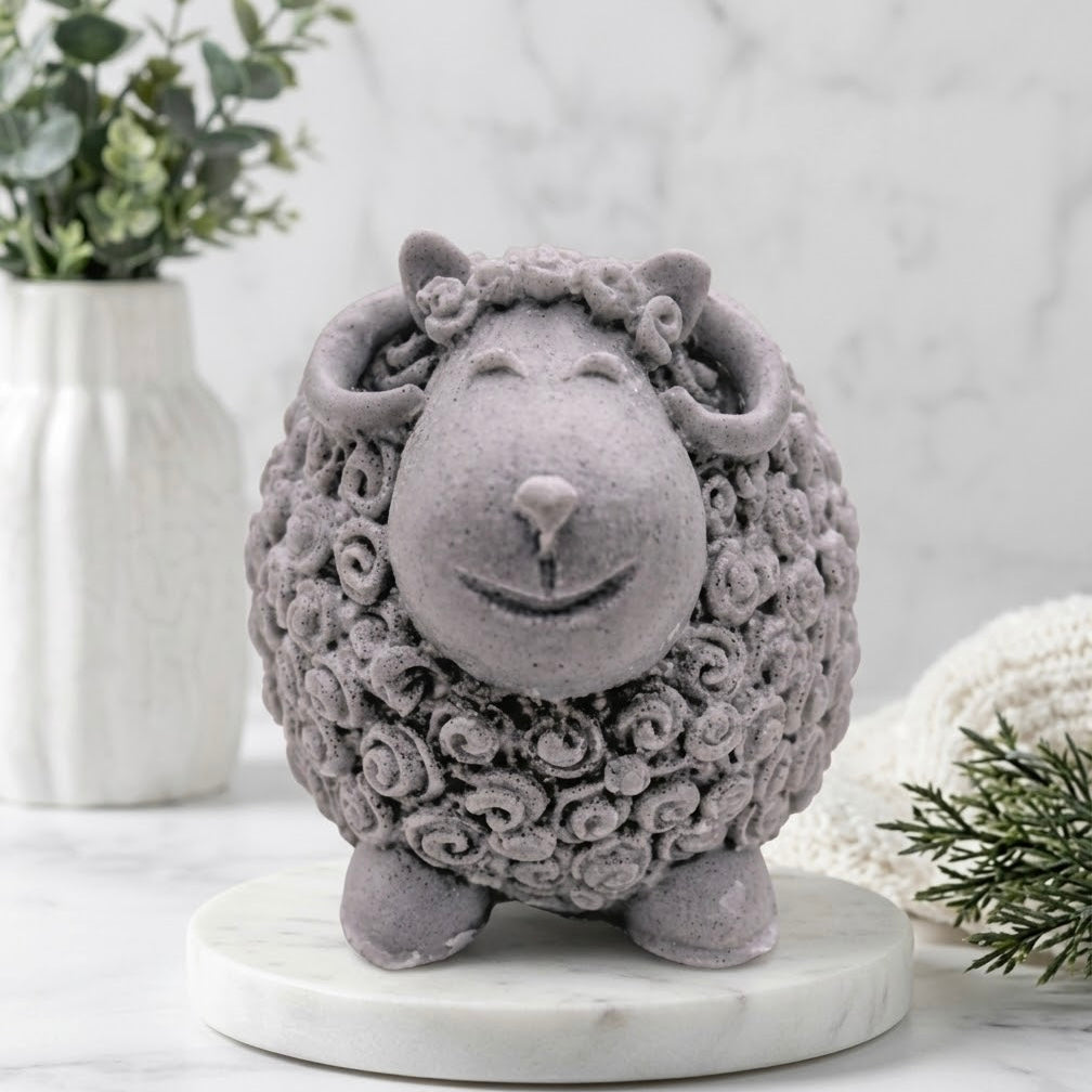 A grey sculpted sheep / ram soap with curly wool texture, standing on a round white marble base. The figurine is styled on a marble surface with a white ceramic vase of greenery and a white knit blanket in the background.