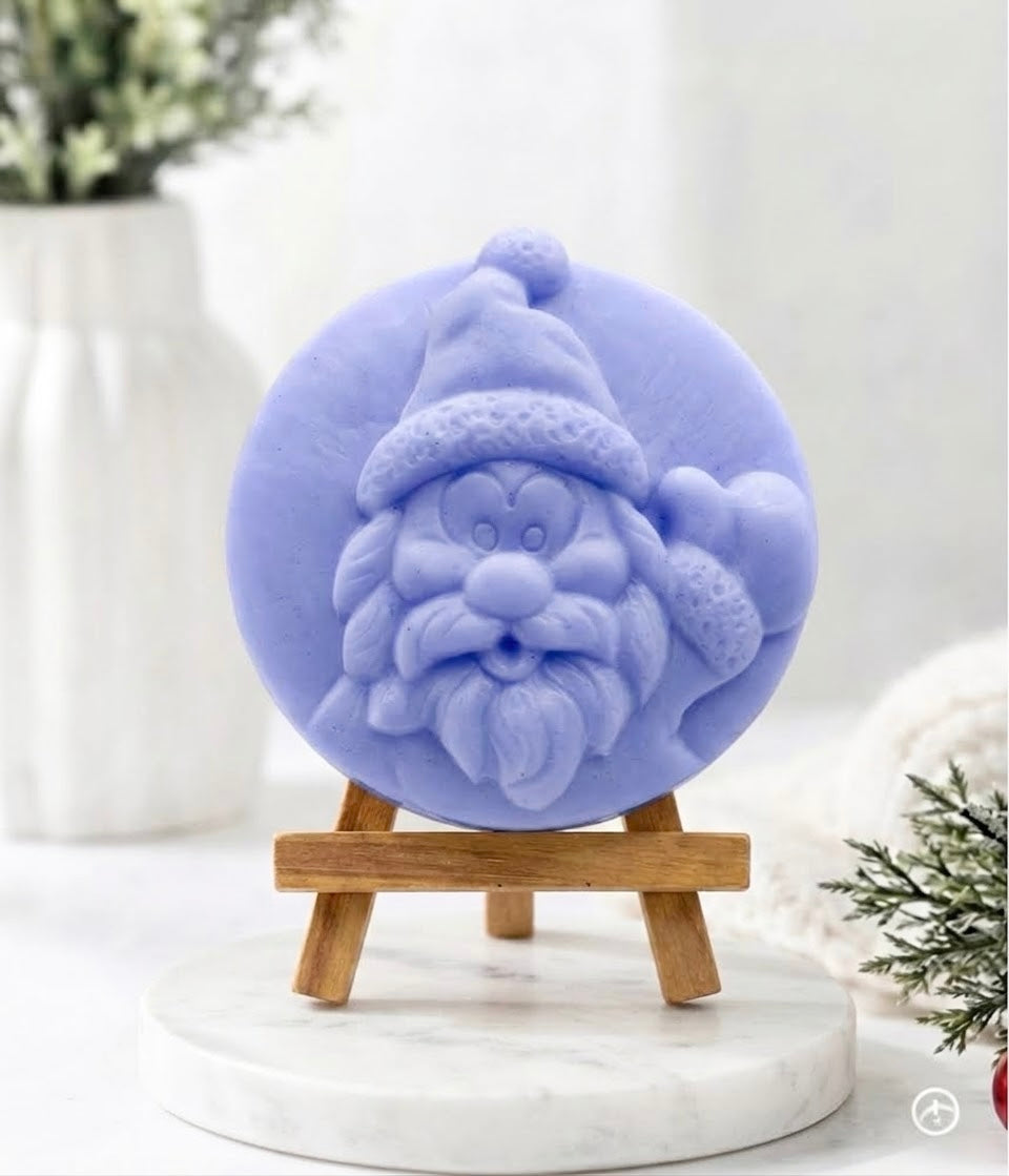 A detailed close-up product photograph of a circular, hand-carved soap disc featuring a relief of Santa Claus’s face, rendered in a distinct periwinkle-lavender-blue colour. The soap disc is displayed on a small wooden tripod easel, which rests upon a white marble pedestal with soft grey veining. The background includes a white fluted vase with blurry green plant sprigs and a soft white knitted textile with a fir branch, creating a cosy and modern holiday feel.