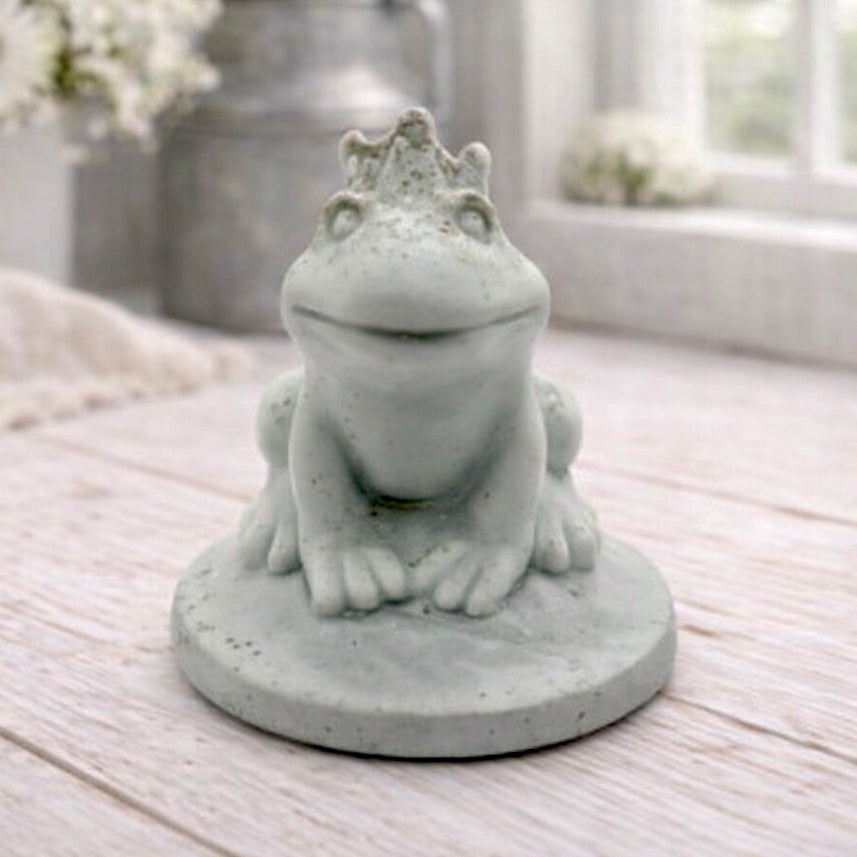 A 3D handmade vegan soap in a pale mint-green tone, sculpted into a sitting frog wearing a crown on its head, positioned on a circular base and surrounded by delicate white flowers in a bright farmhouse setting.