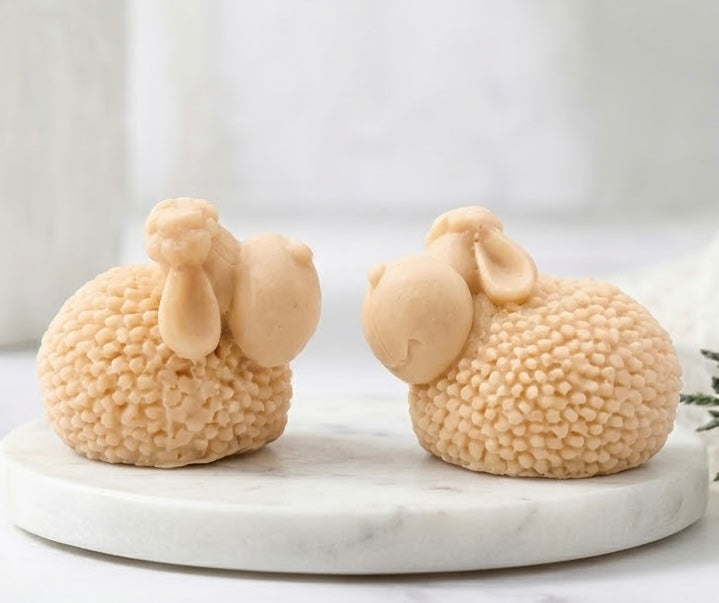 A side profile view of the two cream-coloured, highly textured sheep-shaped soaps with long ears, showcasing the bumpy wool detail, resting on a marble trivet.