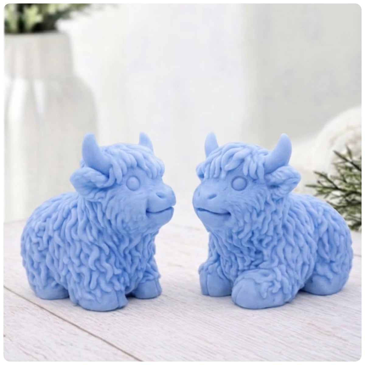 Two blue Highland cow soaps made using indigo powder, are sitting side-by-side and facing each other on a wooden surface, with a blurry background of a vase and a towel