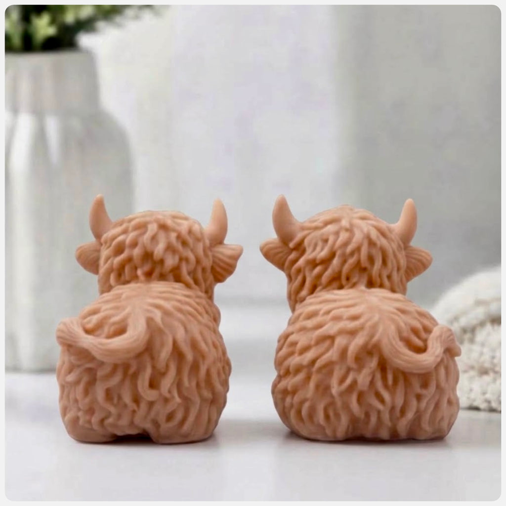Rear view of the two red French clay Highland cow vegan soaps, sitting side-by-side, showing their intricately shaggy fur texture and curled tails from behind.