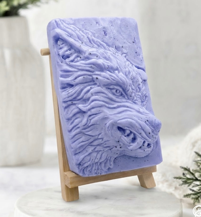 A blue rectangular vegan soap bar featuring a raised relief carving of a wolf's head, displayed on a small wooden easel stand against a soft, blurred background.