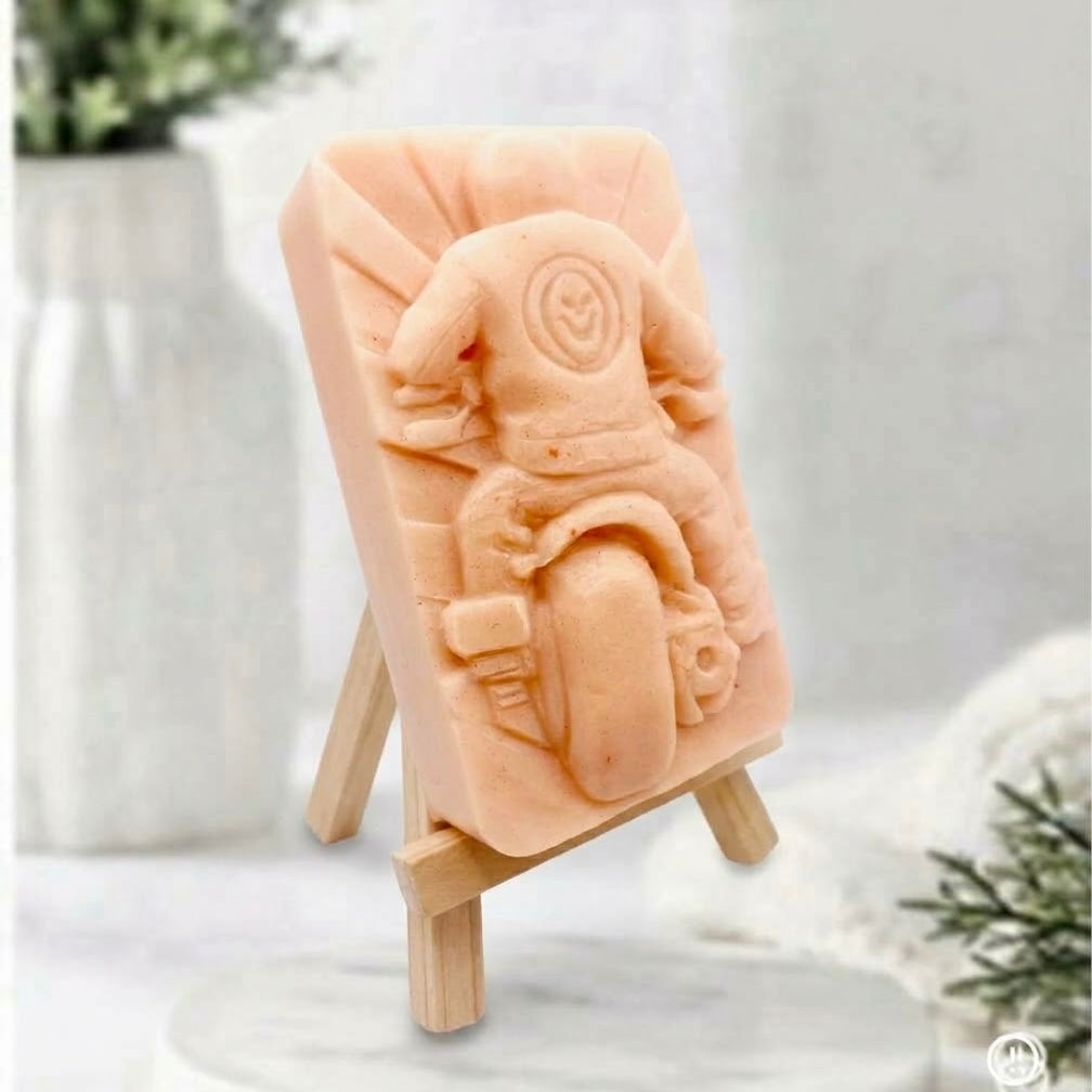 A side view of a pink decorative motorbike soap with a detailed carving of a rider from behind and skull motif, displayed on a wooden stand upon a marble pedestal.