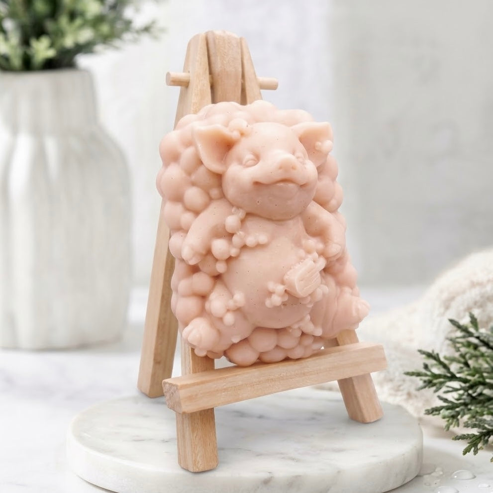 A three-quarter angle view of a pale pink, intricately pig-shaped soap figurine with a bubble-cluster base, resting on a miniature wooden easel, showcasing the detailed textures of the soap.