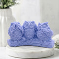 A lavender blue soap sculpture of three owls perched on a branch, displayed on a round white marble pedestal with a blurred white vase in the background.