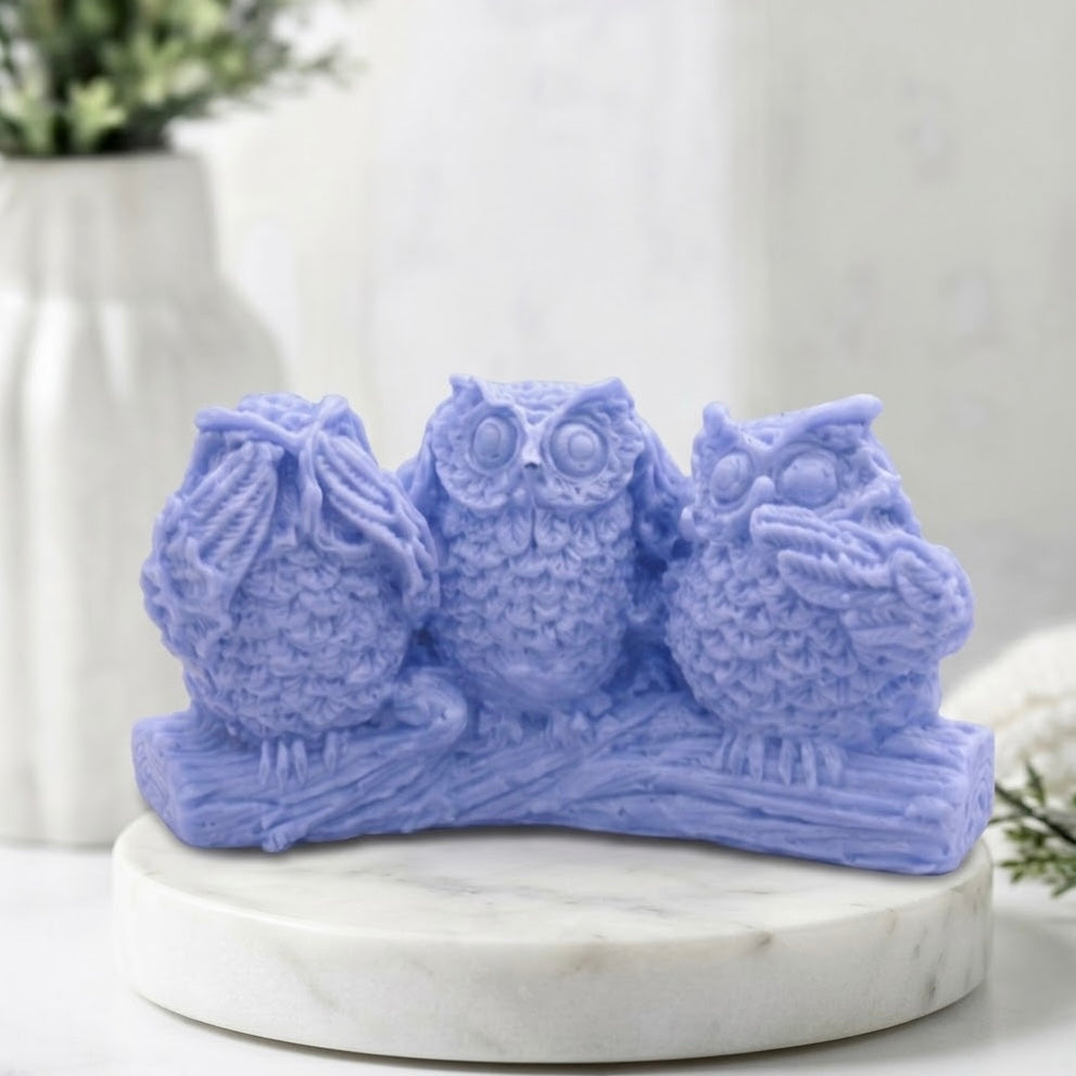 A lavender blue soap sculpture of three owls perched on a branch, displayed on a round white marble pedestal with a blurred white vase in the background.