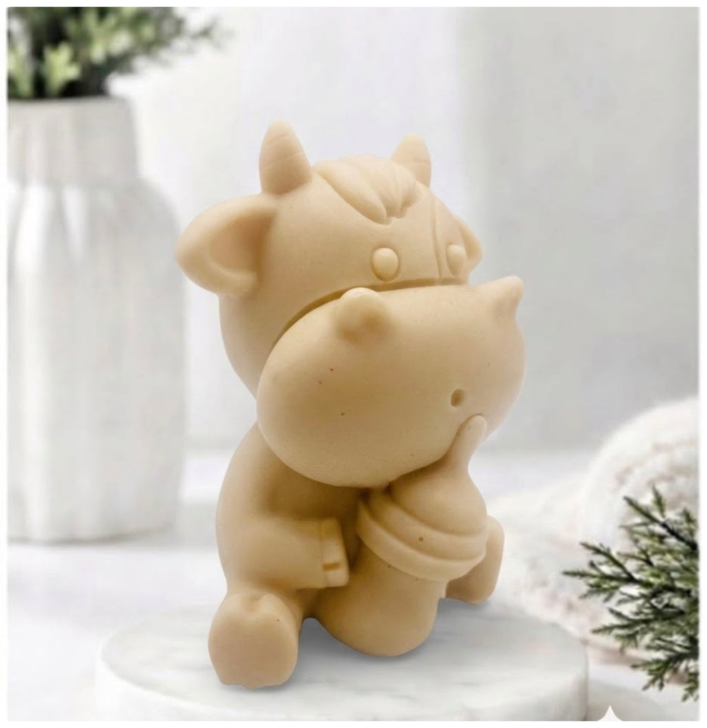 A baby cow-shaped soap holding a small milk bottle, photographed on a soft white background with a blurred bottle and greenery in the surroundings.