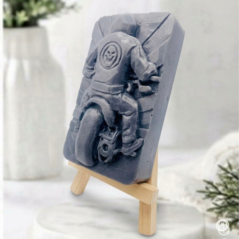 A side view of a charcoal gray handmade motorbike soap featuring a detailed relief carving of a motorcycle rider and skull logo, standing on a mini wooden easel.