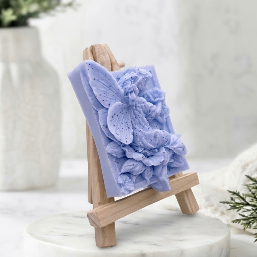A lavender-coloured soap bar in the shape of a butterfly with layered flower petals, displayed on a small wooden easel stand.