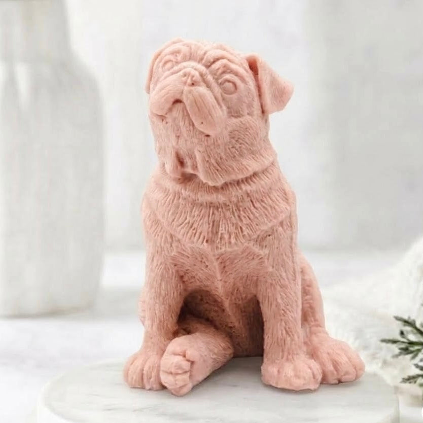 A front, full-body view of a sitting, sculpted pug dog soap in a light pink colour. The soap rests on a round marble pedestal in a tranquil spa environment, with a vase of greenery in the background.