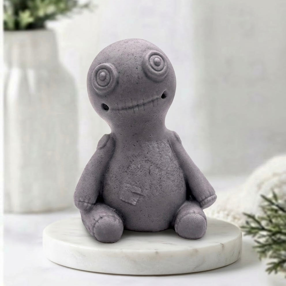 This charcoal grey voodoo doll soap is highly detailed, including spiral eyes, a stitched mouth, and carved heart and belly patches, against a bright, softly blurred bathroom background