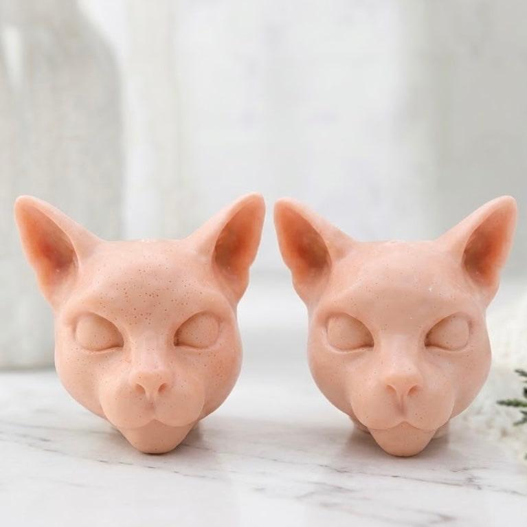 A close-up view of two handmade vegan soap bars in a light pink colour, detailed with the features of Siamese cat heads, resting side-by-side on a white marble vanity.