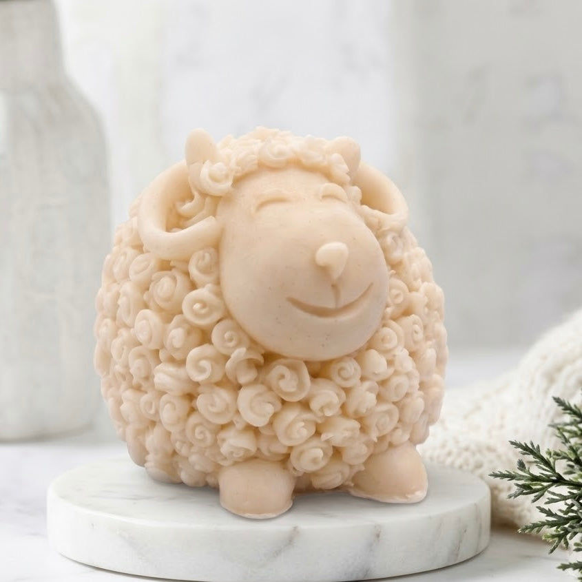 A cute, smiling sheep soap with curly cream-coloured wool, standing on a round white marble base. The background is a soft, blurred white marble surface with a white textured vase and a small sprig of greenery.