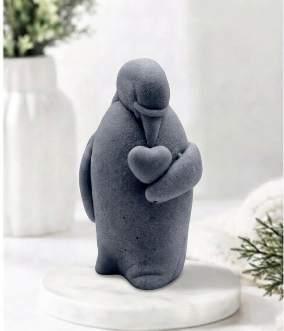 Artisan grey textured penguin soap figurine holding a heart, standing on a round white marble display stand with a soft-focus background of a fluted vase and festive greenery.