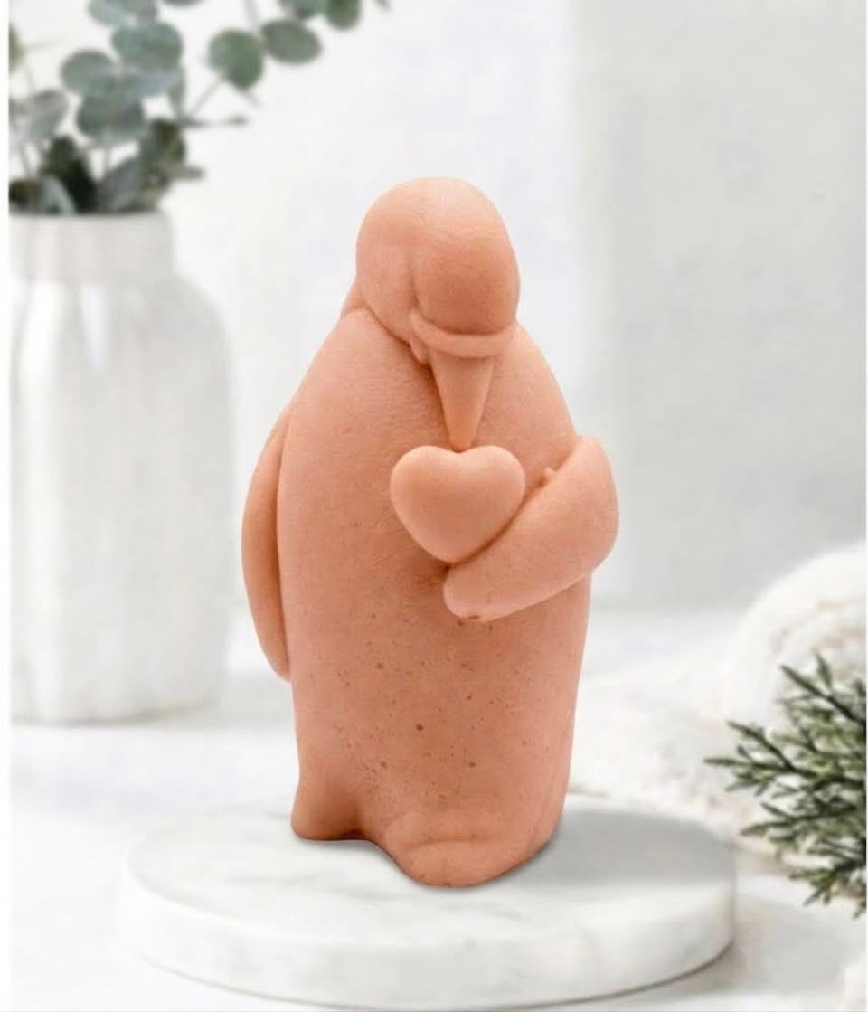 Artisan terracotta-coloured penguin soap sculpture holding a matching heart, standing on a white marble display stand in a beautifully lit, cosy interior setting.