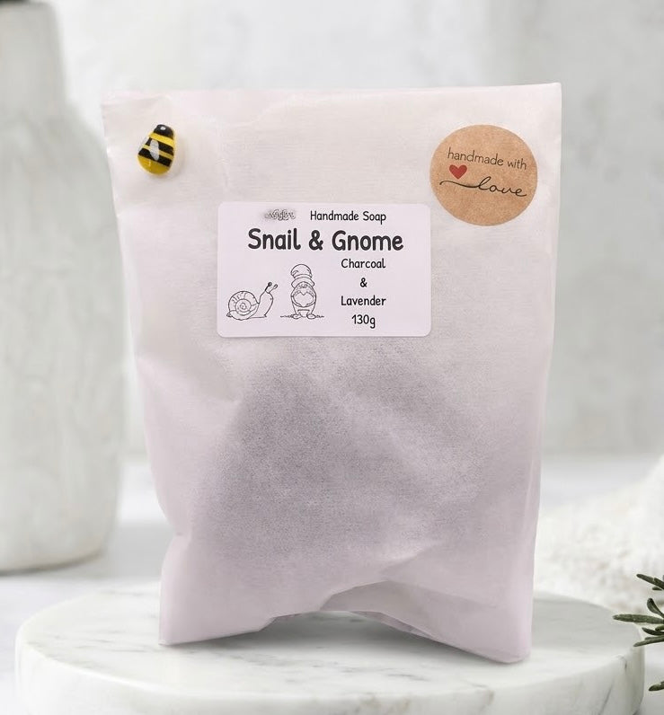 A 130g bag of Snail & Gnome handmade soap, charcoal & lavender, shown on a white marble pedestal with a soft-focus background.