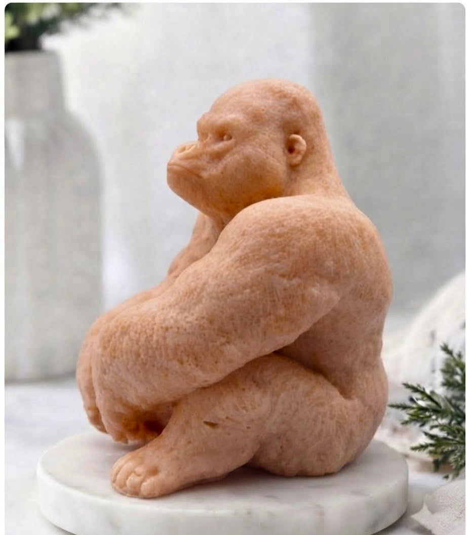 A sculpted vegan soap of a seated gorilla in a warm red French clay tone, displayed on a round white marble base against a softly blurred indoor background.