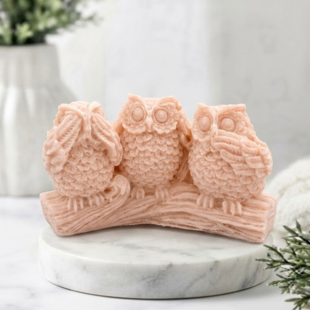 A pink soap wise owl figurine sitting on a branch, displayed on a round white marble pedestal with a softly blurred indoor background.