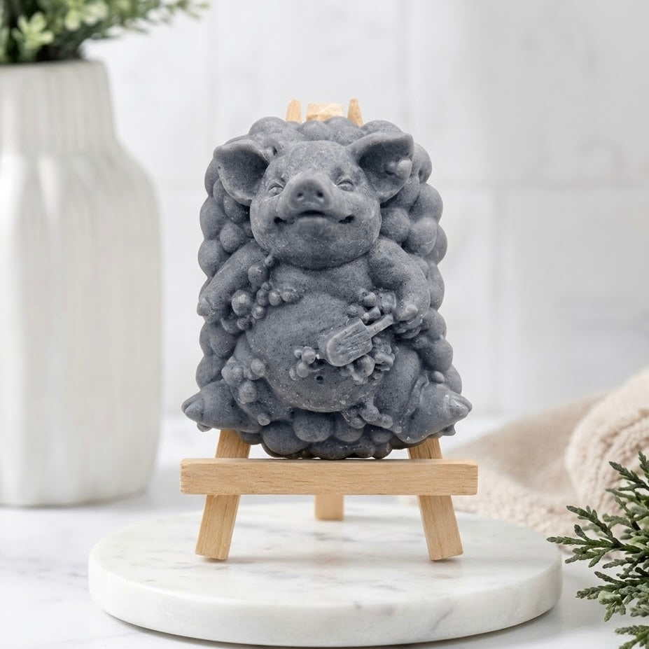 Full-frontal view of a pig-shaped soap figurine with a bubble-cluster base, made in a charcoal grey colour, resting full-frontal on a miniature wooden easel.
